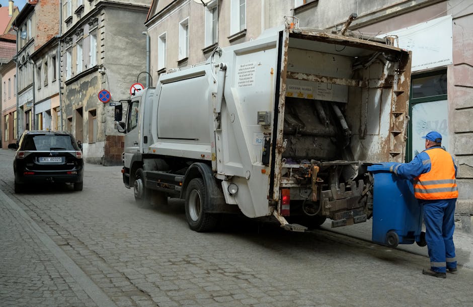 A white waste collection truck with a large, open rear hopper positioned on a narrow cobblestone street next to an old, multi-storey building with beige and gray facades. The truck shows signs of usage, with rust and dirt marks visible inside the rear compartment. To the right, a worker dressed in a high-visibility orange vest, blue uniform, and a blue cap is emptying a blue wheeled bin into the truck’s hopper. The street also has parked cars, one black vehicle directly behind the truck, with a license plate visible. The scene takes place during daylight, with natural light illuminating the environment, and the surrounding buildings exhibit a mix of classical architectural elements with decorative window frames and worn paintwork. The activity appears to be part of an on-site rubbish removal process, relevant for alternative waste disposal methods managed by companies like Rubbish Removal Ilford, operating within urban environments requiring private waste collection services.