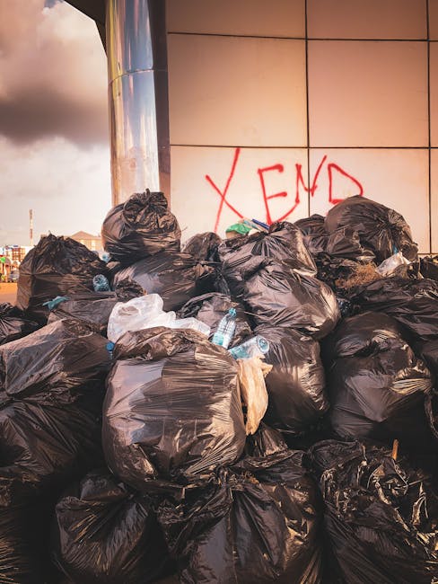 Three black plastic rubbish bags, filled and tied at the top, are placed on the curbside on a street, positioned next to a low wall and a black metal fence. The bags appear to contain household waste and are slightly crumpled, with some areas showing creases and folds on their surface. Behind the fence, dense foliage of bushy trees and shrubs can be seen, creating a textured green backdrop. The scene is illuminated by natural daylight, casting soft shadows on the pavement, and the environment suggests a typical residential area where private waste disposal or rubbish removal services, such as those provided by Rubbish Removal Ilford, might be used for on-site clearance or alternative rubbish collection outside regular council collection times.