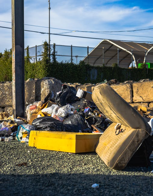 A pile of mixed household rubbish is gathered on a gravel surface outside, consisting of black plastic garbage bags, some partially torn open revealing various waste contents. Among the debris, a yellow plastic container and a large, dismantled or discarded beige car tyre are prominently visible, with the tyre leaning to the right and partially overlapping the rubbish. Behind the pile, there is a low stone wall constructed from irregularly shaped stones, with some green foliage peeking over the top. In the background, a metal fence and trees are visible, along with a structure that has a curved, metallic roof, suggesting a storage or industrial area. Overhead, power lines stretch across a partly cloudy sky, indicating a semi-urban environment suitable for private waste collection and rubbish removal services, such as those provided by Rubbish Removal Ilford for local waste management or alternative disposal methods. The scene is lit by natural daylight, creating clear shadows and highlighting the textures and colors of the waste materials and surroundings.
