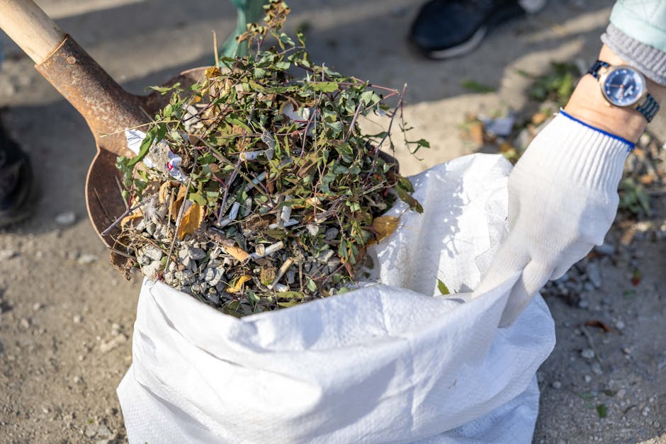 A pile of mixed household rubbish is gathered on a gravel surface outside, consisting of black plastic garbage bags, some partially torn open revealing various waste contents. Among the debris, a yellow plastic container and a large, dismantled or discarded beige car tyre are prominently visible, with the tyre leaning to the right and partially overlapping the rubbish. Behind the pile, there is a low stone wall constructed from irregularly shaped stones, with some green foliage peeking over the top. In the background, a metal fence and trees are visible, along with a structure that has a curved, metallic roof, suggesting a storage or industrial area. Overhead, power lines stretch across a partly cloudy sky, indicating a semi-urban environment suitable for private waste collection and rubbish removal services, such as those provided by Rubbish Removal Ilford for local waste management or alternative disposal methods. The scene is lit by natural daylight, creating clear shadows and highlighting the textures and colors of the waste materials and surroundings.