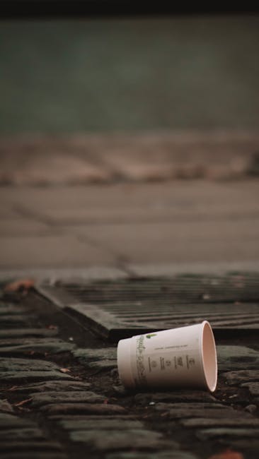 A white disposable coffee cup with a lid is lying on its side on a paved surface made of brick or stone, with some dirt and debris visible nearby. The cup appears to be empty and is positioned close to a metal drainage grate or vent embedded in the pavement. In the background, there is a blurred, greenish outdoor environment, possibly grass or foliage, contributing to an overall impression of an urban or suburban street scene. The lighting is natural but somewhat subdued, suggesting an overcast day. This scene reflects typical instances where waste, such as discarded cups, can be found on pavements or public pathways, a common concern addressed by private rubbish removal services like those offered by Rubbish Removal Ilford, which specialise in clearing waste from various outdoor or internal spaces to keep areas clean and free from litter.