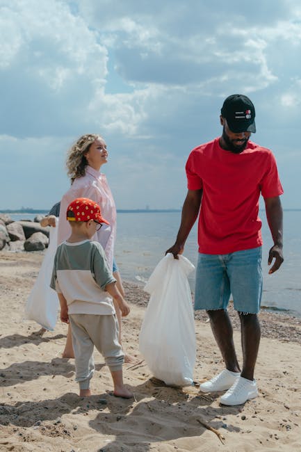 A man in a red t-shirt, denim shorts, white sneakers, and a black cap is standing on a sandy beach, holding a large white rubbish bag. Beside him, a young boy wearing a red hat with a pattern, light grey sweatpants, and a grey and white t-shirt is also holding a white rubbish bag. In the background, a woman with curly blonde hair, wearing a light pink dress, is facing the man and boy, engaged in the activity. The scene is set on a beach with a shoreline visible, featuring large rocks along the water's edge. The sky above is partly cloudy with some sunlight filtering through, creating a bright but soft atmosphere. The overall scene suggests outdoor rubbish collection or cleanup activity, subtly related to waste management and environmental preservation, aligning with the focus of rubbish removal services through a natural, real-life context.