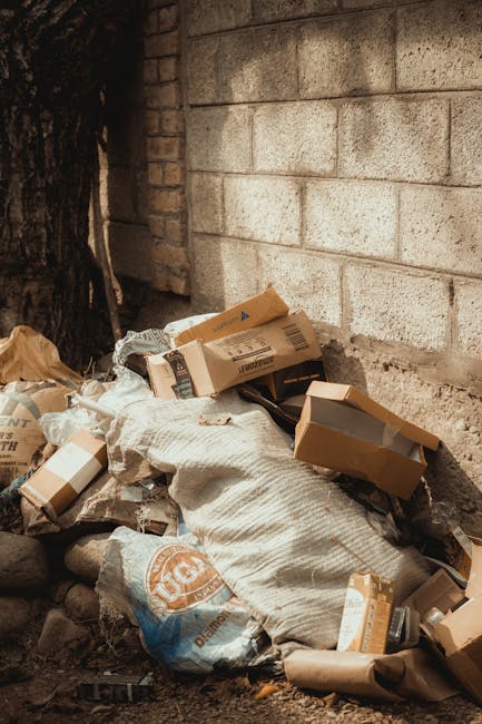 A collection of discarded cardboard boxes and packaging materials are piled on the ground in front of a brick wall with visible mortar joints, adjacent to a tree trunk on the left side of the image. The boxes, some partially collapsed or torn, vary in size and are made of brown corrugated cardboard. Among the waste, there are plastic bags and fabric-like sacks, one of which bears a faded logo. The ground beneath the rubbish consists of dirt and small stones, indicating an outdoor setting, possibly a back alley or a garden corner. Natural daylight illuminates the scene with soft shadows cast by the objects and nearby structures. This image exemplifies a typical scenario where private waste disposal through an independent rubbish removal service, such as Rubbish Removal Ilford, might be needed to manage such clutter effectively, especially when local authority collection is delayed or unavailable.