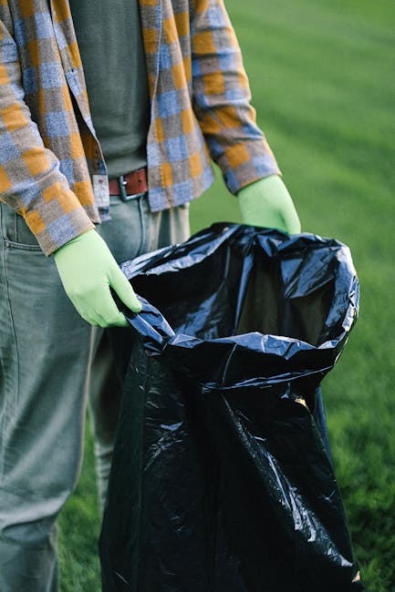 A person wearing a yellow and grey checkered shirt, grey trousers, and light green gloves is holding open a large black garbage bag, which is lined with a shiny black plastic inner layer. The individual is standing outdoors on a grassy area with vibrant green grass visible in the background, suggesting an environment suitable for rubbish collection or waste disposal. The scene appears to be part of an on-site waste removal process, potentially related to private rubbish removal services in Ilford. The trash bag is empty but prepared for rubbish collection, indicating active engagement in rubbish or waste management, similar to alternative waste handling solutions offered by companies like Rubbish Removal Ilford. The natural outdoor lighting emphasizes clear visibility of the materials and textures, with the focus on the task of rubbish collection, supporting the context of environmentally responsible waste disposal practices.