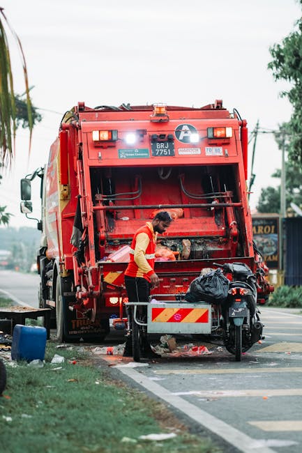 A worker wearing a yellow high-visibility vest and black trousers operates a red rubbish collection vehicle, positioned on the side of a street. The vehicle's rear compartment is open, revealing mechanical components and empty space for debris. The worker is seen handling black waste bags placed on a small metal platform attached to the vehicle. Nearby, a black motorbike with a black bag secured on its seat is parked close to the waste collection vehicle. The street setting includes a paved curb, some scattered debris, and a grassy verge with a blue plastic container lying on its side. In the background, there are trees, utility poles, and a few commercial signs, indicating a suburban or urban area. The scene is during the daytime with natural lighting, reflecting a typical rubbish removal activity possibly conducted by a private waste management service like Rubbish Removal Ilford, in line with alternative waste collection methods for local residents.