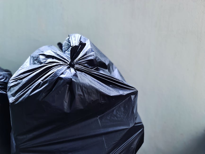 A close-up view of a large black plastic rubbish bag made from shiny, crinkled material, tightly knotted at the top, positioned in the foreground against a plain, light-colored wall. The bag appears filled with waste, with creases and folds visible on its surface, and is likely placed for collection or disposal as part of a rubbish removal service. In the background, partially visible, are additional black bags, suggesting an accumulation of refuse typical for domestic or small-scale commercial waste collection, possibly carried out by a private waste handling service like Rubbish Removal Ilford. The setting is an indoor or outdoor area with minimal environmental details, focusing on the bags which are ready to be moved or disposed of, aligning with the context of rubbish collection and waste management.
