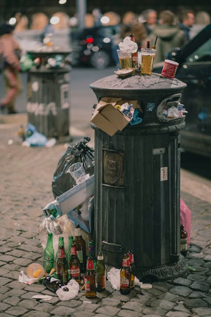 A black, cylindrical public rubbish bin made of metal with a domed lid, positioned on a cobblestone pavement. The bin is overflowing with various types of waste, including paper, plastic, and glass bottles, with some trash spilling onto the ground around its base. On top of the lid are several disposable paper coffee cups, some with plastic lids, and a few paper food containers, one of which is open with a visible food item inside. The surrounding area includes a background with blurred pedestrians walking, parked cars, and additional rubbish scattered on the street, indicating a busy urban environment. The scene emphasizes the importance of waste management and the potential for rubbish to accumulate when proper disposal practices are not followed, aligning with the themes of independent waste collection and rubbish removal services such as those offered by Rubbish Removal Ilford.