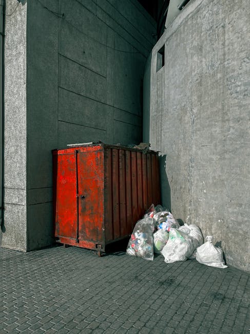 A weathered, large red metal skip with visible rust and chipped paint is positioned in an outdoor urban environment, placed against the junction of a dark grey building and a light grey concrete wall. The skip is rectangular with vertical ribbing on its sides and a closed top, situated on a patterned, dark paved surface. Adjacent to the skip, several clear plastic rubbish bags filled with mixed waste are piled at the base, some slightly leaning against the container. The background shows the corner of a modern building with smooth, dark grey cladding and a small square window near the top of the concrete wall. The lighting appears natural and diffused, typical of an overcast day, highlighting the textures of the metal, concrete, and waste materials. This scene exemplifies a typical setup for on-site waste collection, possibly managed by private rubbish removal services, illustrating the practical aspects of independent rubbish disposal options relevant for residents and businesses in the area.
