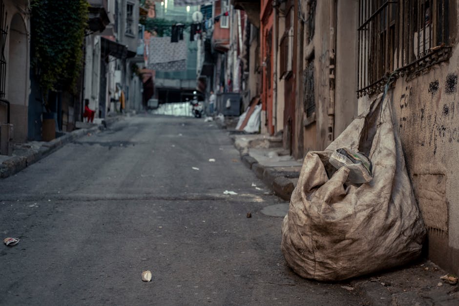A large, dirty, beige fabric sack filled with discarded waste materials is situated on the right side of an uneven urban street, positioned directly against a weathered brick wall with metal security bars on the windows. The sack appears worn and stained from prolonged exposure to the outdoor environment. Surrounding the sack, small pieces of litter such as paper and plastic debris are scattered on the asphalt surface of the street, which has visible cracks and patches. In the background, additional residential buildings with varied facades and laundry hanging on lines between them can be seen, indicating an inner-city environment. The scene is illuminated by natural daylight, casting soft shadows on the street, and the overall atmosphere suggests a location where private or informal waste disposal occurs, potentially illustrating an area with rubbish access issues aligning with independent rubbish removal solutions. Rubbish Removal Ilford's services could be relevant here for addressing accumulated debris and improving environmental cleanliness in such settings.