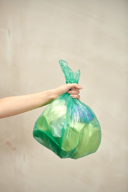 A person's hand is shown holding a tied green plastic trash bag, which appears full and slightly bulging. The bag is made of thin, semi-translucent plastic with visible creases and folds, and contains miscellaneous waste materials, giving it a rounded shape. The person grips the bag firmly with their fingers wrapped around the gathered plastic, with the thumb visible underneath. The background is a plain, neutral-toned wall, providing no specific environmental context. The lighting is soft and even, highlighting the glossy surface of the plastic bag without harsh shadows. This scene reflects a typical example of waste collection or rubbish removal, relevant to services offered by companies like Rubbish Removal Ilford, which specialise in quick and efficient waste clearance solutions, including private disposal and on-site rubbish removal at various premises.