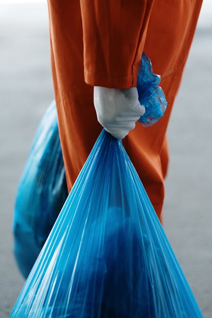 Close-up of a person wearing an orange protective suit and white gloves holding a blue plastic rubbish bag by its gathered top, the bag filled and slightly bulging, with additional rubbish bags visible in the background, suggesting an ongoing waste collection or clearance activity. The setting appears to be outdoors, possibly on a paved surface or near a waste disposal area. The scene emphasizes the handling of waste materials in a professional and sanitary manner, aligning with rubbish removal services focused on efficient and safe disposal. The image reflects the process of private waste handling, highlighting the importance of proper protective equipment and available waste bags for on-site rubbish collection, consistent with services offered by Rubbish Removal Ilford for urgent or same-day waste clearance.